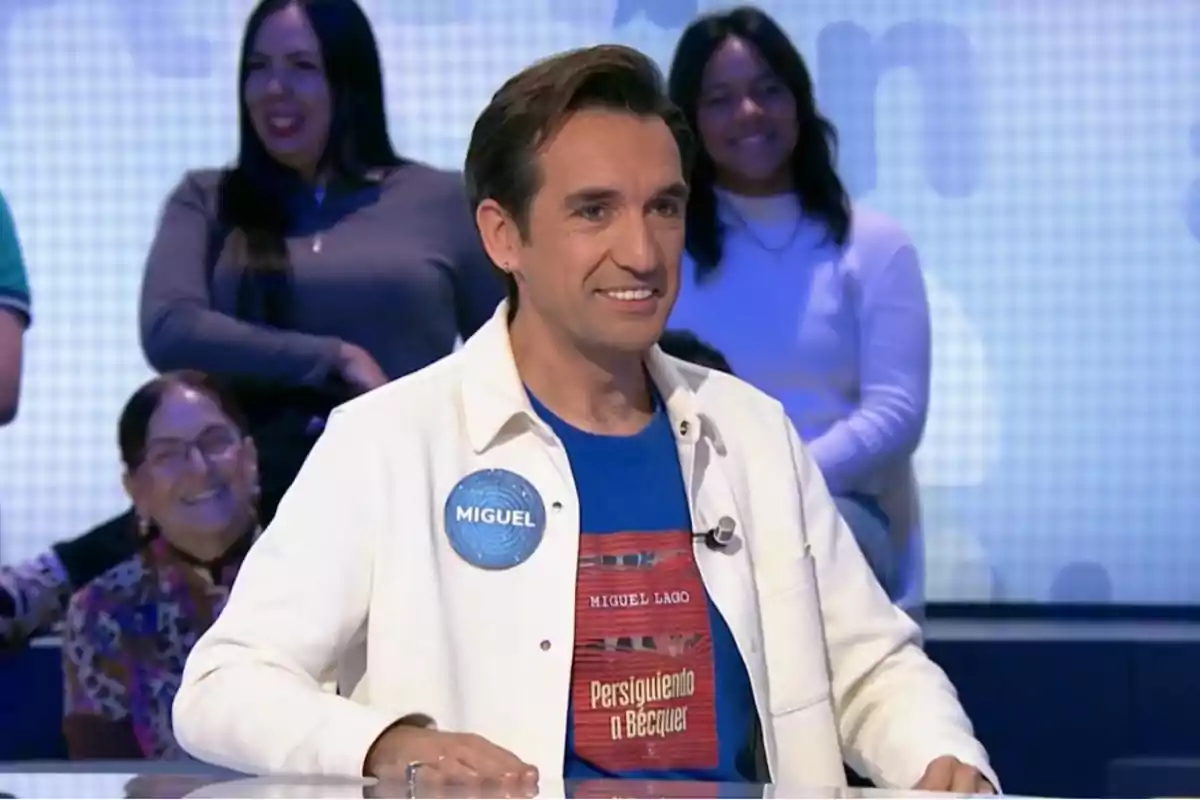 Miguel Lago con una camisa blanca y una camiseta azul sonríe mientras está sentado en un programa de televisión con personas en el fondo.