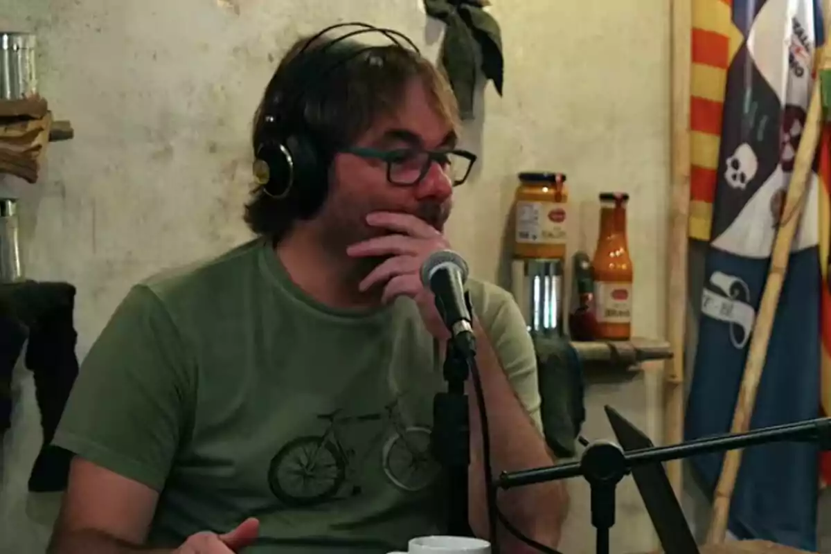 Quim Masferrer con gafas y auriculares está sentado frente a un micrófono en un entorno de grabación, con una camiseta verde que tiene una bicicleta dibujada. Quim Masferrer con gafas y auriculares está sentado frente a un micrófono en un entorno de grabación, con una camiseta verde que tiene una bicicleta dibujada.