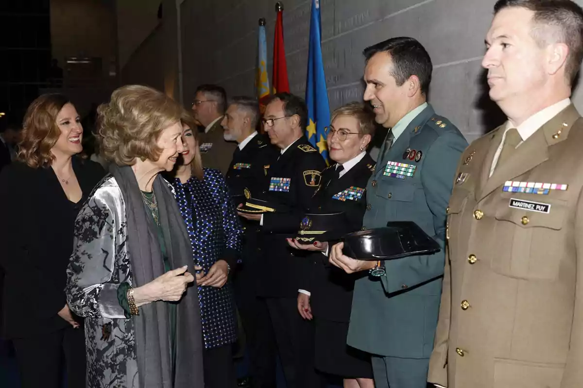 Un grupo de personas, algunas en uniforme militar, están de pie en fila en un evento formal, mientras la reina Sofía interactúa con ellos. Un grupo de personas, algunas en uniforme militar, están de pie en fila en un evento formal, mientras la reina Sofía interactúa con ellos.