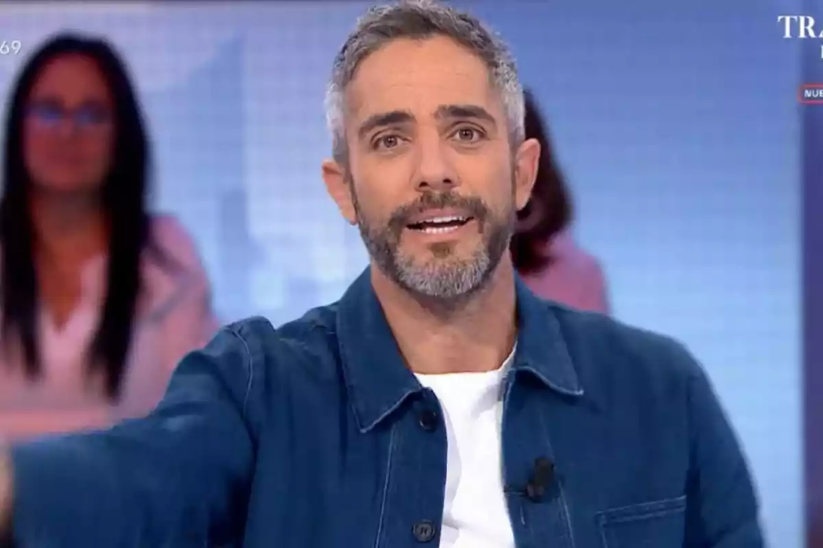 Roberto Leal con barba y cabello corto canoso, vistiendo una chaqueta azul y camiseta blanca, está hablando en un estudio de televisión con un fondo desenfocado.