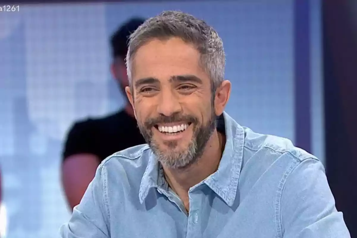 Roberto Leal sonriente con barba y camisa de mezclilla en un estudio de televisión.