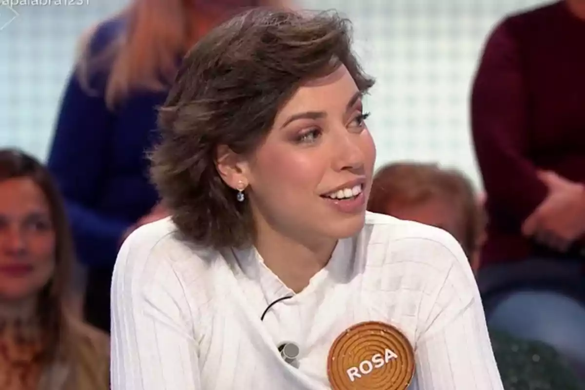 Rosa con cabello corto y castaño sonríe mientras participa en un programa de televisión, lleva un suéter blanco y un broche con el nombre 