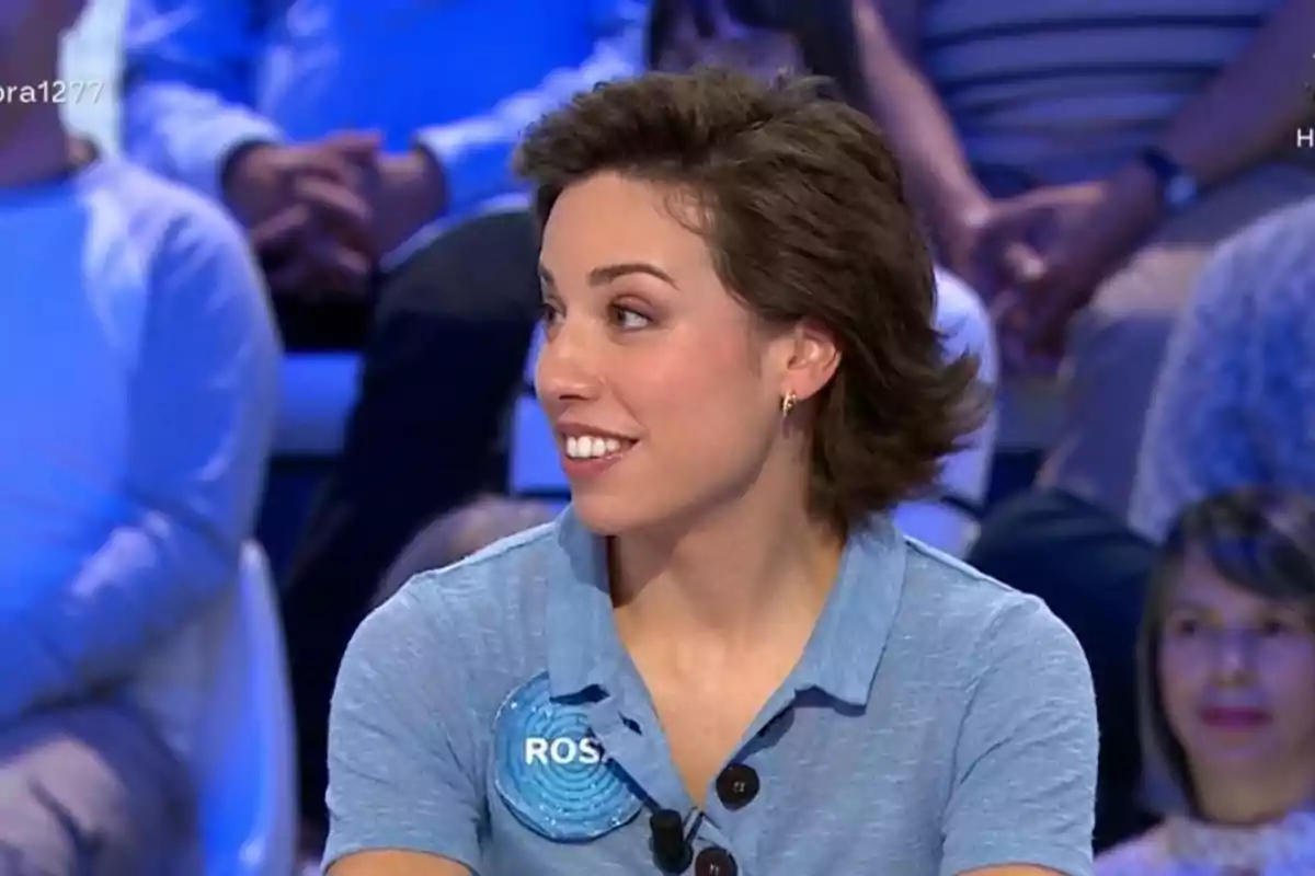 Rosa con cabello corto y castaño, vestida con camisa azul claro y una placa con el nombre "ROS", sentada en un plató de televisión con público al fondo