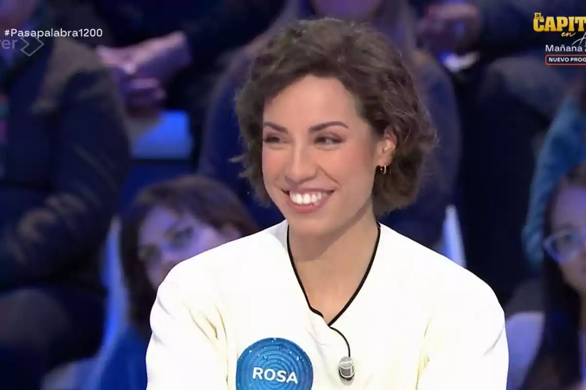 Rosa sonriente con cabello corto y rizado, lleva una camiseta blanca con un micrófono de solapa y un círculo azul con el nombre Rosa sonriente con cabello corto y rizado, lleva una camiseta blanca con un micrófono de solapa y un círculo azul con el nombre