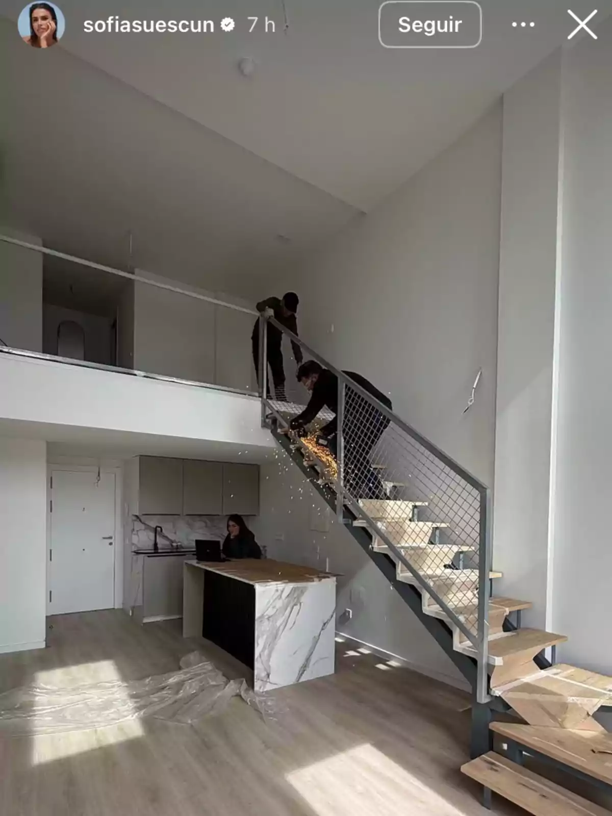 Una persona trabaja en una cocina moderna mientras dos personas instalan una barandilla en una escalera de madera en un espacio de concepto abierto. Una persona trabaja en una cocina moderna mientras dos personas instalan una barandilla en una escalera de madera en un espacio de concepto abierto.