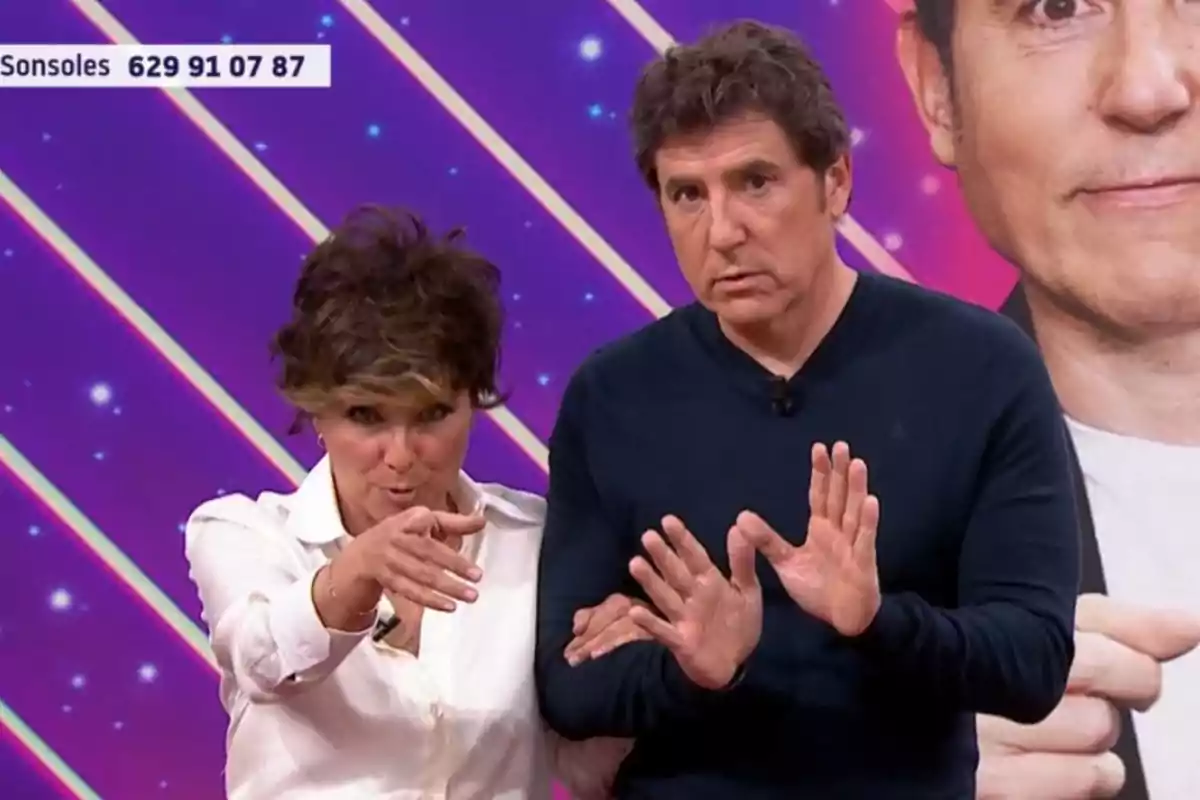 Sonsoles Ónega y Manel Fuentes en un set de televisión con un fondo colorido y un número de teléfono visible en la esquina superior izquierda. Sonsoles Ónega y Manel Fuentes en un set de televisión con un fondo colorido y un número de teléfono visible en la esquina superior izquierda.