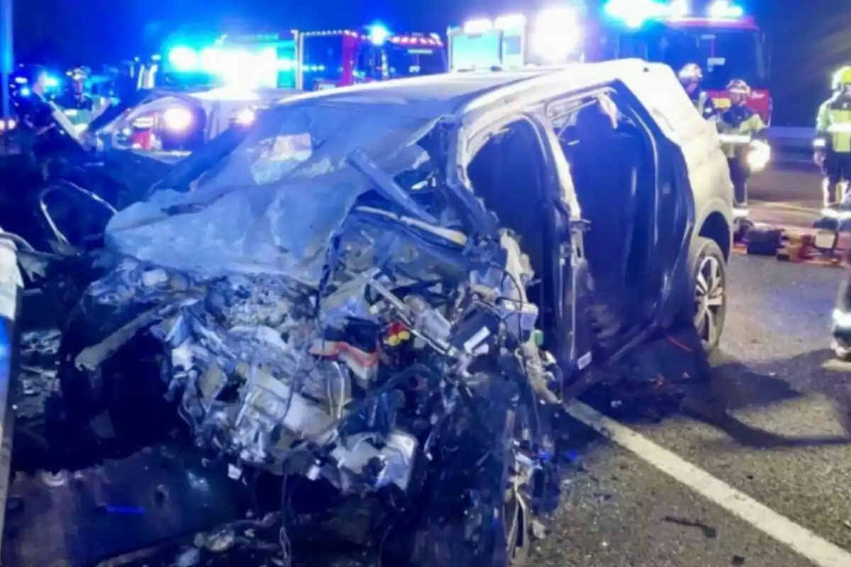 Auto gravemente dañado tras un accidente en carretera de noche con luces de emergencia y personal de rescate al fondo