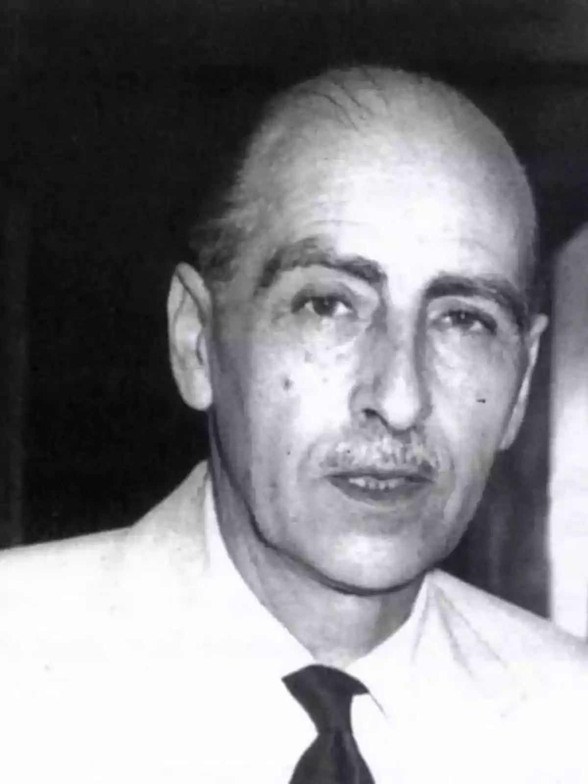 Hombre de mediana edad con bigote y cabello corto, vestido con camisa clara y corbata oscura, mirando hacia la cámara en una fotografía en blanco y negro Hombre de mediana edad con bigote y cabello corto, vestido con camisa clara y corbata oscura, mirando hacia la cámara en una fotografía en blanco y negro