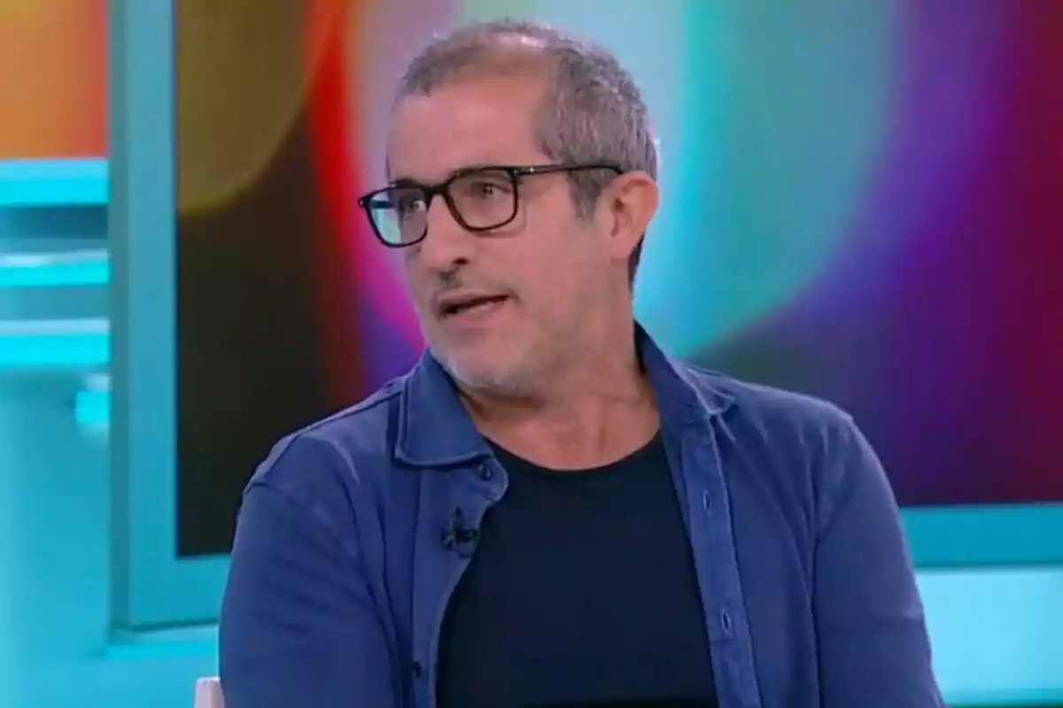 Un hombre con gafas y barba canosa hablando en un programa de televisión con un fondo colorido.