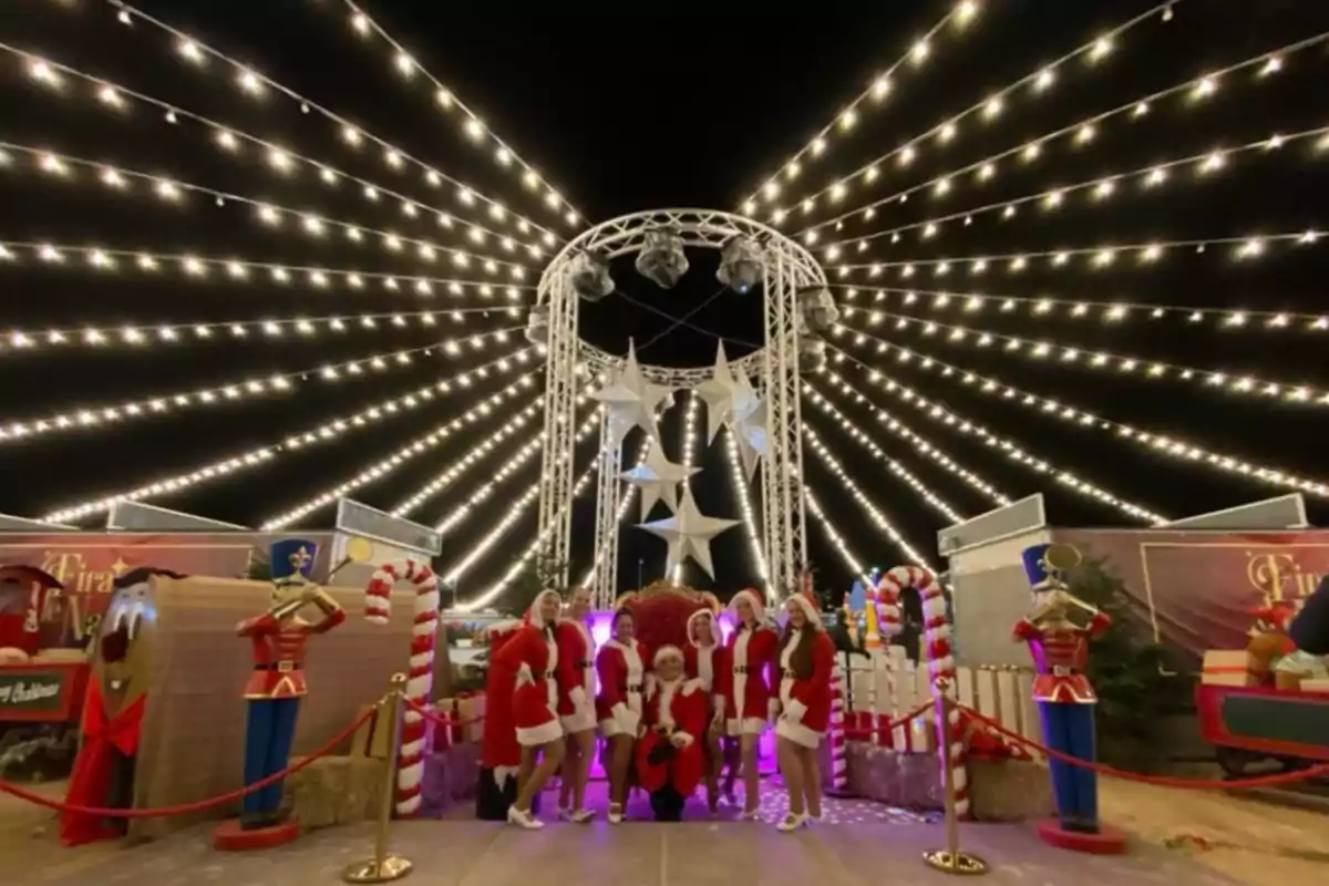 Grupo de personas vestidas de Santa Claus posando en un escenario navideño iluminado con luces y decoraciones festivas.