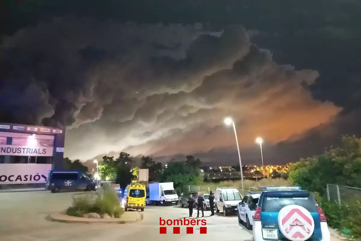 Una escena nocturna muestra una gran nube de humo oscuro sobre un área industrial iluminada por luces de emergencia y vehículos de servicios de emergencia estacionados en el lugar.