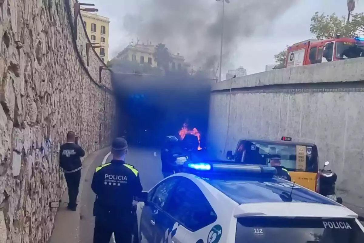 Imagen ed un coche incendiado en la Ronda Litoral de Barcelona mientras agentes de la Guardia Urbana intervienen en el incidente