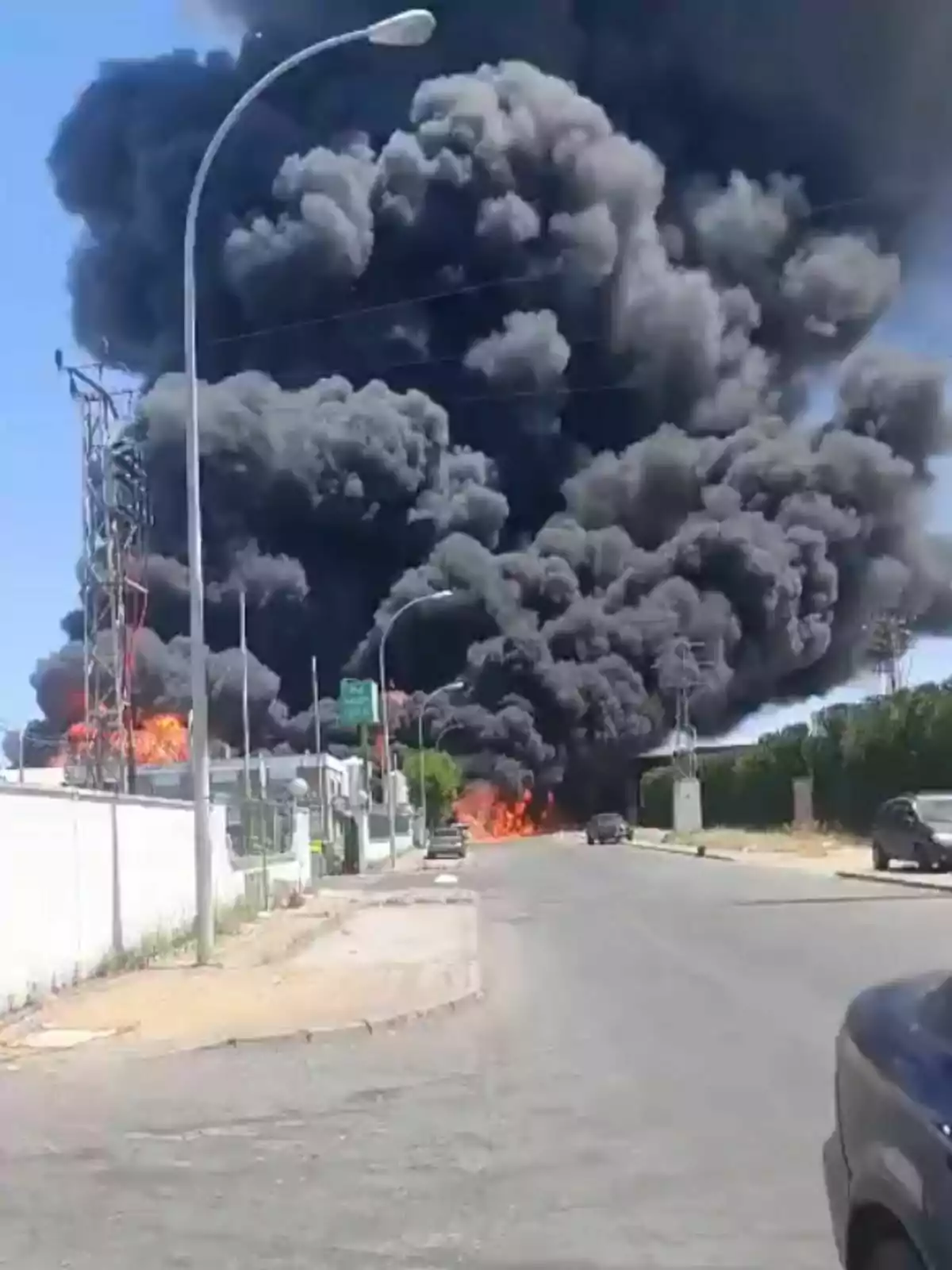 Una gran columna de humo negro se eleva desde un incendio en una zona urbana de Sevilla, con llamas visibles y vehículos estacionados en la calle cercana. Una gran columna de humo negro se eleva desde un incendio en una zona urbana de Sevilla, con llamas visibles y vehículos estacionados en la calle cercana.
