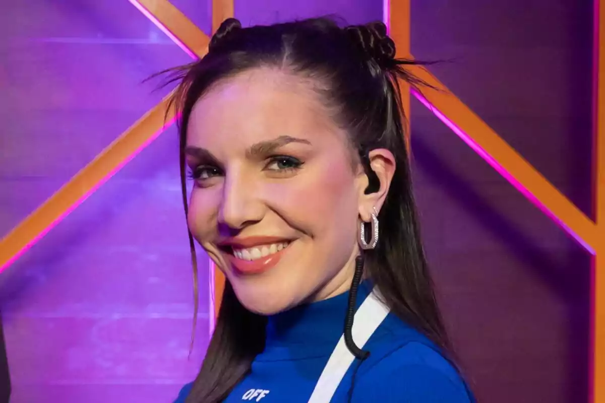 Inés Hernand sonriente con el cabello largo y oscuro, usando un suéter azul y aretes grandes, con un fondo iluminado en tonos morados y naranjas en MasterChef Celebrity.