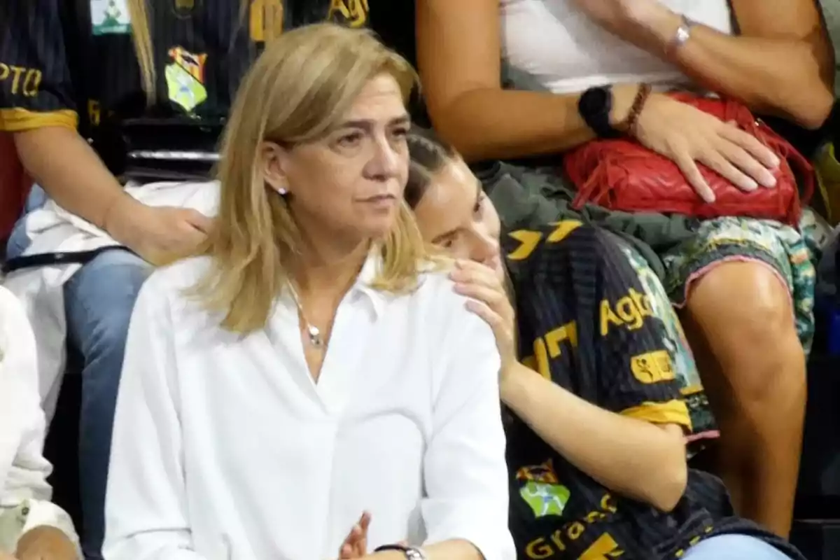 La infanta Cristina de cabello rubio y camisa blanca está sentada en un evento deportivo, con personas alrededor vistiendo camisetas negras con detalles amarillos. La infanta Cristina de cabello rubio y camisa blanca está sentada en un evento deportivo, con personas alrededor vistiendo camisetas negras con detalles amarillos.