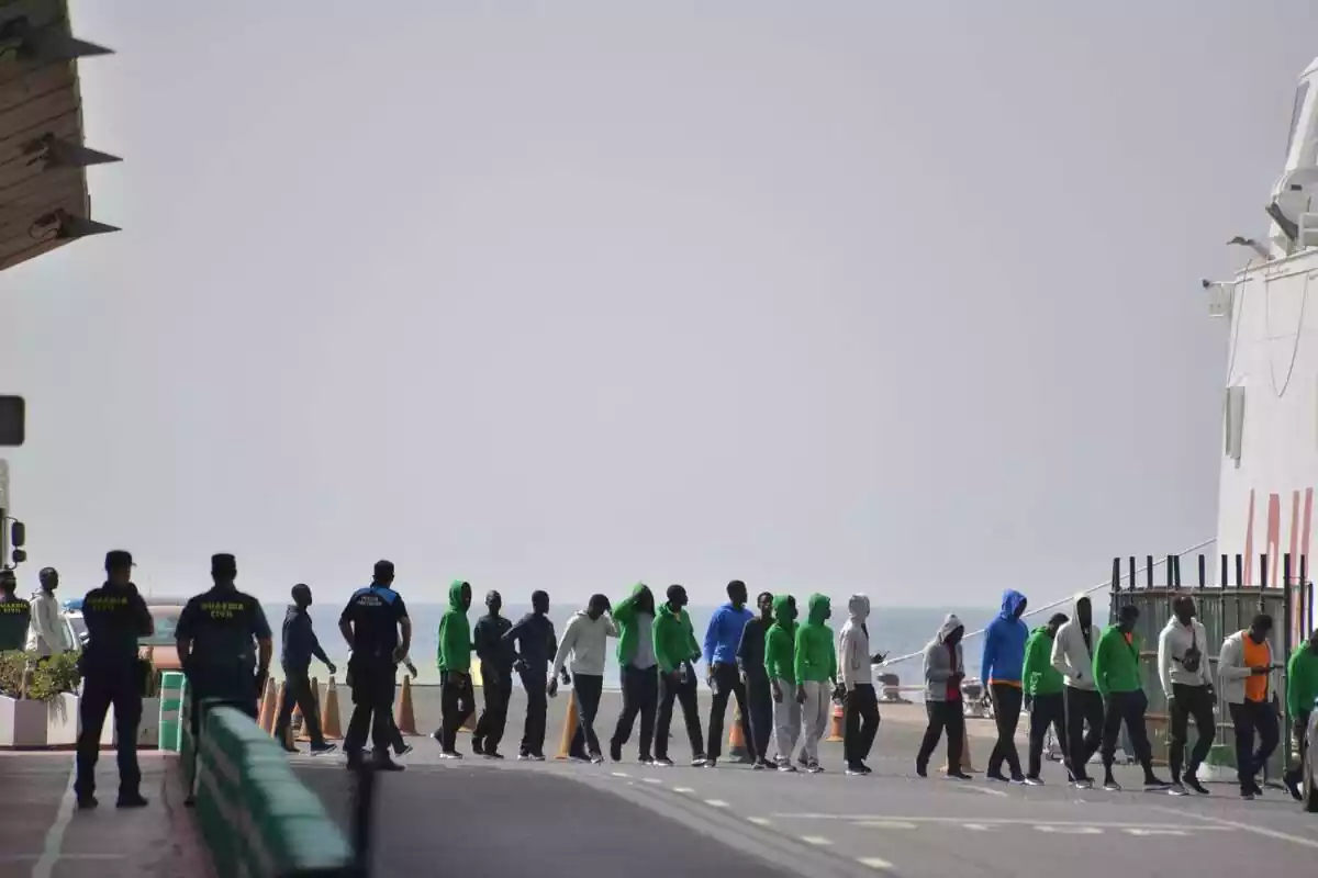 Plano general con una cola de inmigrantes recién llegados a El Hierro subiendo a un barco mientras unos Guardia Civiles los vigilan