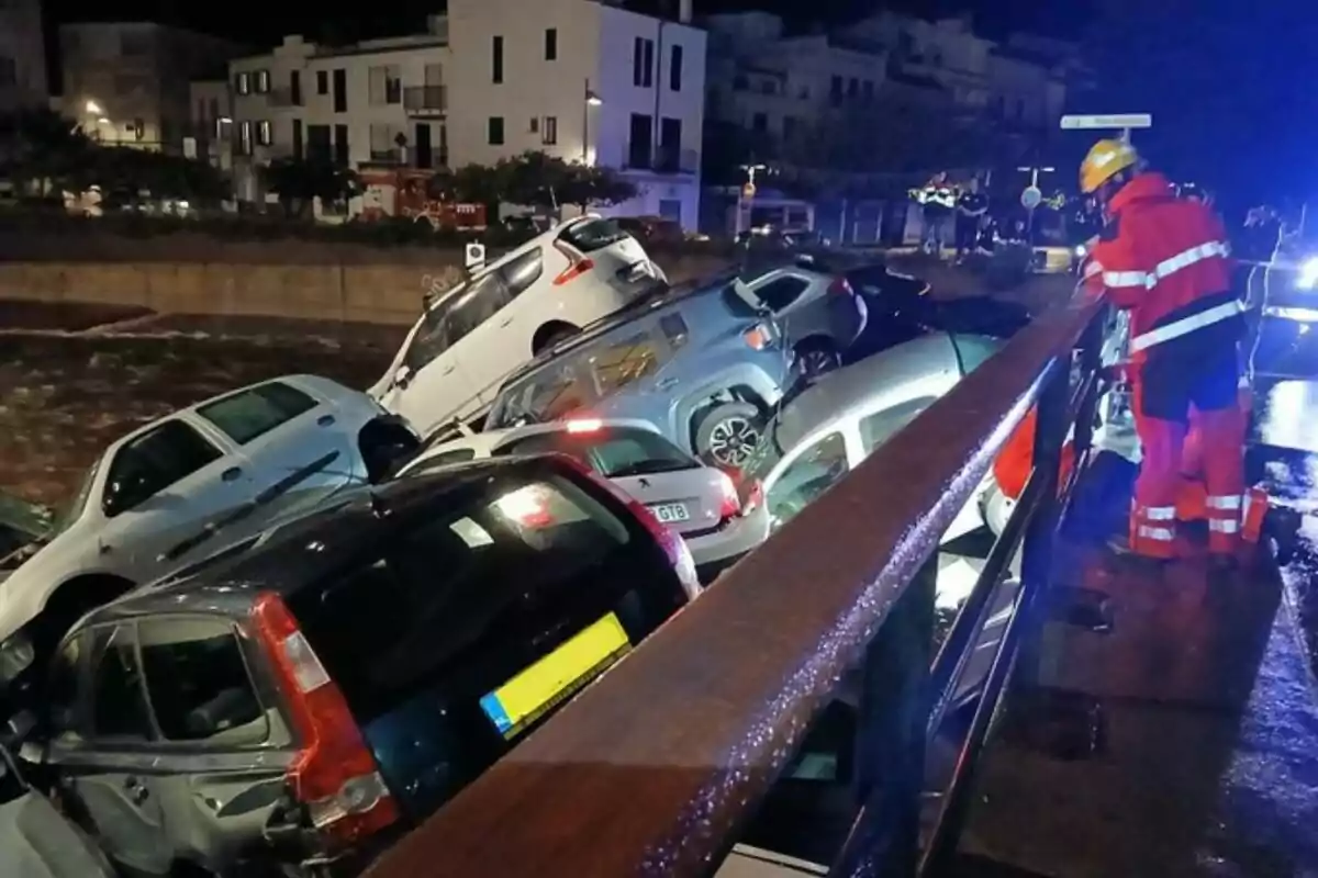 Coches apilados en una calle inundada mientras un rescatista con uniforme rojo observa desde un puente.