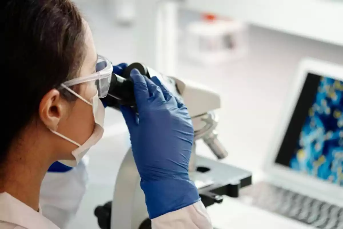 Científica observando a través de un microscopio en un laboratorio con guantes y mascarilla.