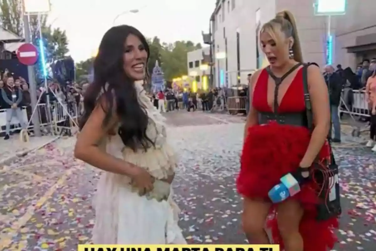 Dos mujeres están en una calle llena de confeti, Isa Pantoja lleva un vestido blanco y Marta Riesco un vestido rojo, mientras un grupo de personas observa desde el fondo en La Familia de la Tele. Dos mujeres están en una calle llena de confeti, Isa Pantoja lleva un vestido blanco y Marta Riesco un vestido rojo, mientras un grupo de personas observa desde el fondo en La Familia de la Tele.
