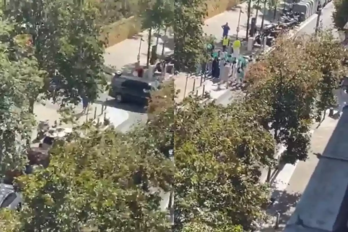 Una imagen aérea muestra una calle arbolada con un grupo de personas reunidas en la acera y un vehículo negro circulando por la vía.