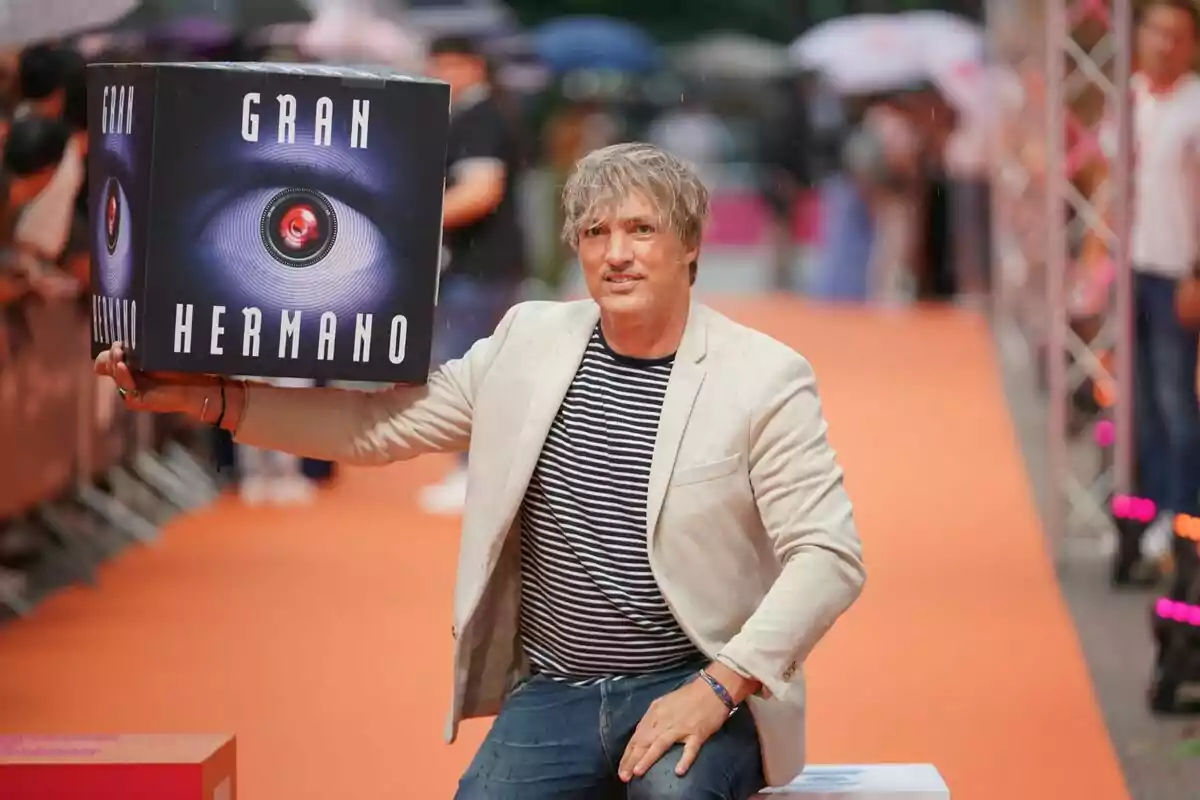 Ismael Beiro con una chaqueta beige y camiseta de rayas sostiene una caja con el logo de "Gran Hermano" en un evento con alfombra naranja.