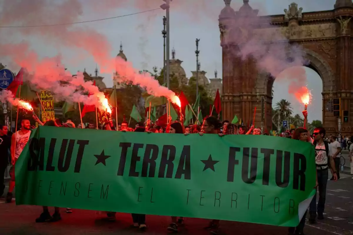 Manifestación con personas sosteniendo bengalas y una pancarta verde que dice "SALUT TERRA FUTUR" frente al Arco de Triunfo.