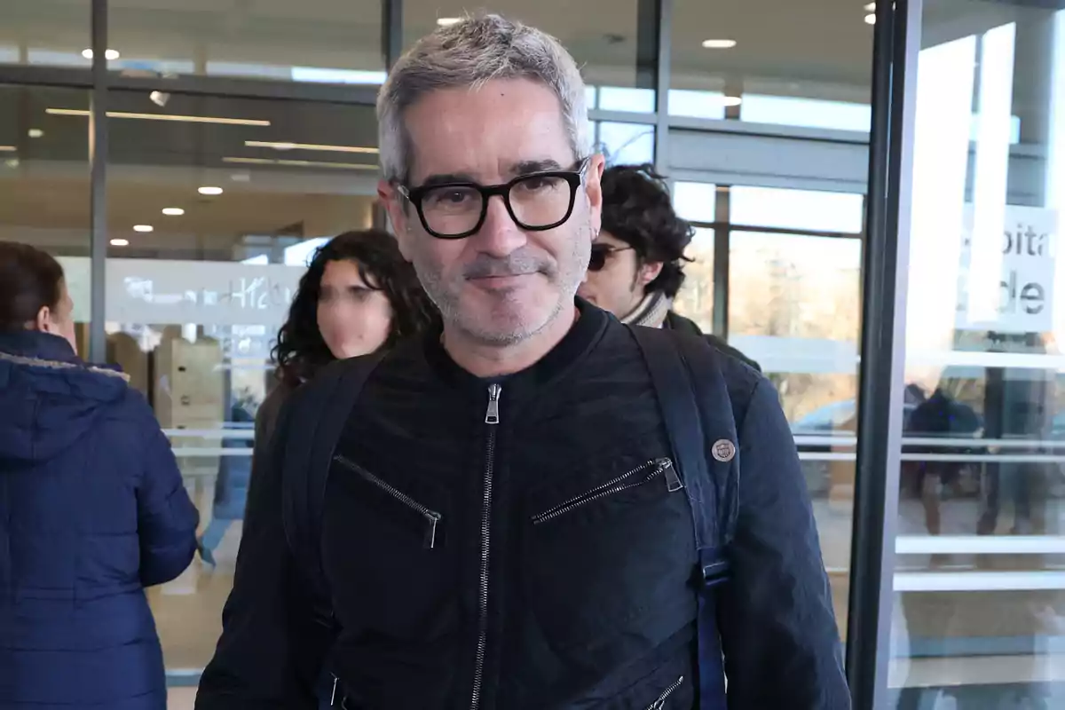 Jacobo Martos con gafas y chaqueta negra está de pie frente a un edificio con puertas de vidrio, rodeado de otras personas. Jacobo Martos con gafas y chaqueta negra está de pie frente a un edificio con puertas de vidrio, rodeado de otras personas.