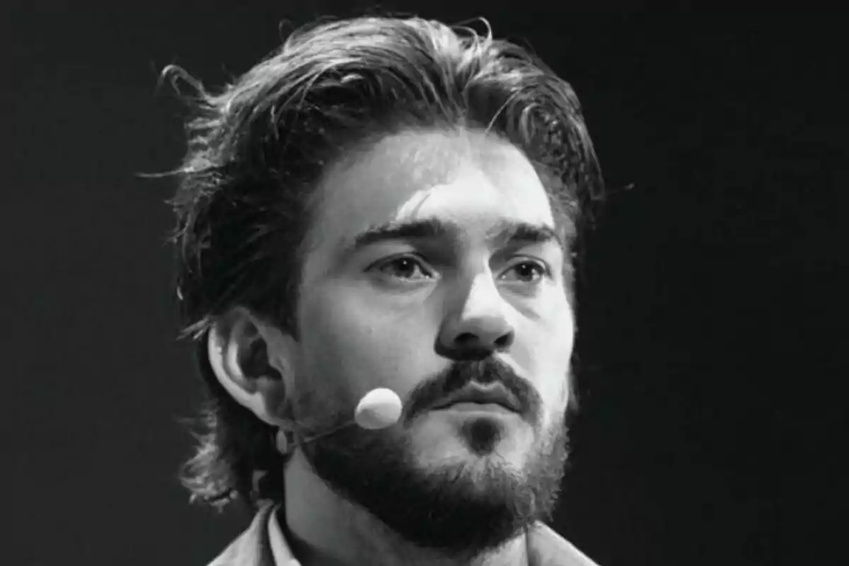 Fotografía en blanco y negro de James Hollcroft con barba y cabello largo usando un micrófono de diadema.