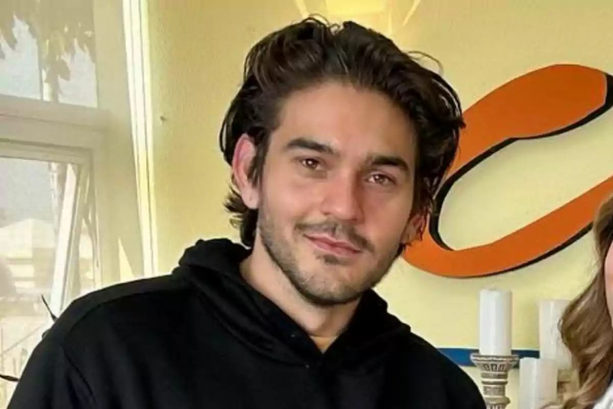 James Hollcroft con cabello oscuro y barba ligera, vistiendo una sudadera negra, posando frente a una pared amarilla con una ventana y una decoración en forma de letra 