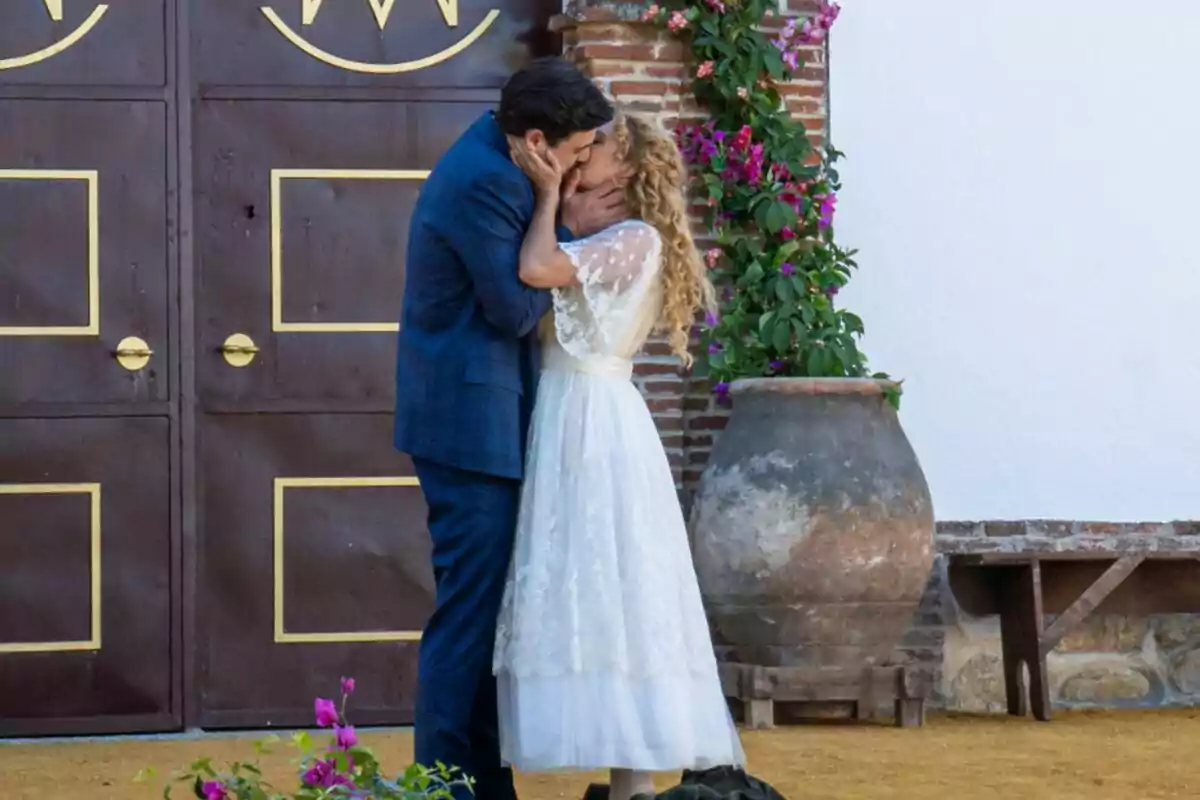 Jana y Manuel se besa apasionadamente frente a una puerta de madera con detalles dorados, junto a una maceta grande y flores en La Promesa.