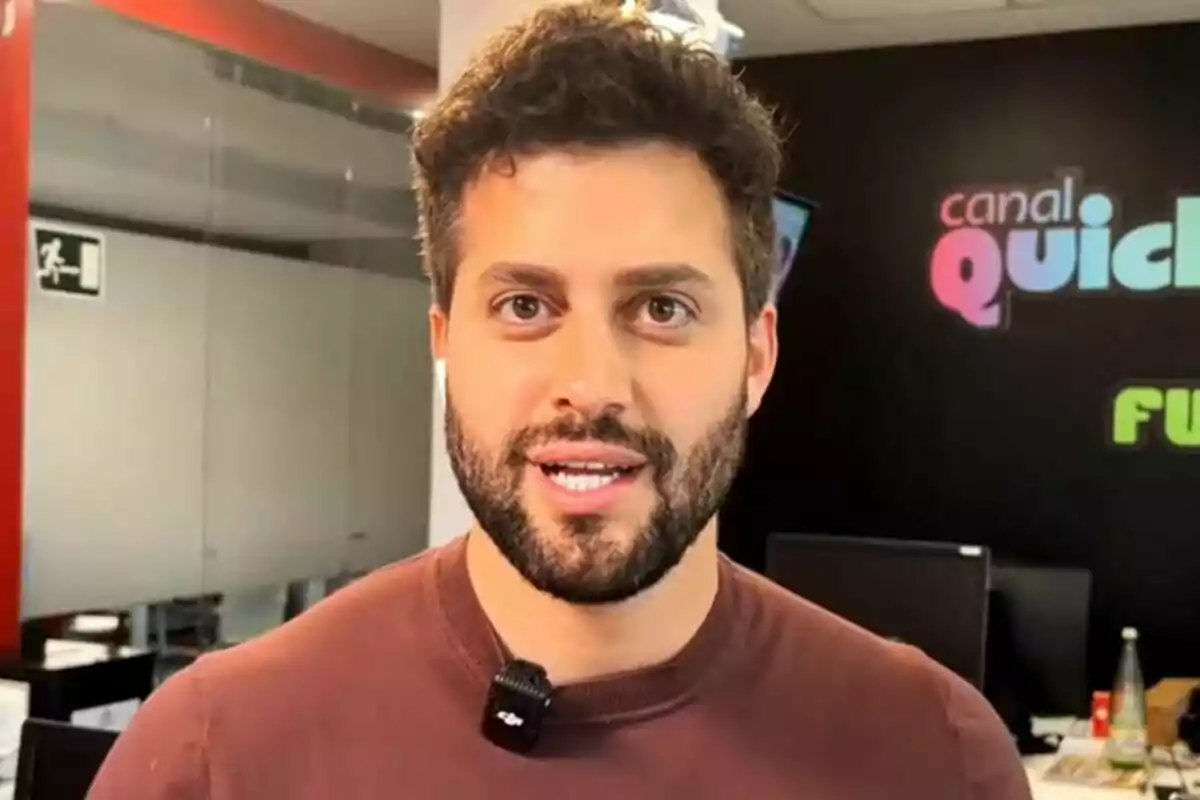 Javi de Hoyos con barba y cabello rizado, usando un micrófono de solapa, está de pie en una oficina con el logo de "canal Quicke" y "Ni que fuéramos" en la pared detrás de él.