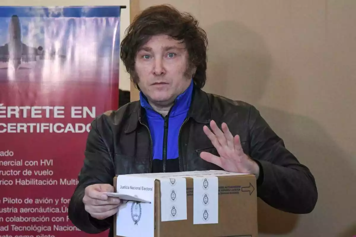 Plano medio de Javier Milei con chaqueta de cuero introduciendo su voto en una urna de cartón y mirando fijamente a cámara