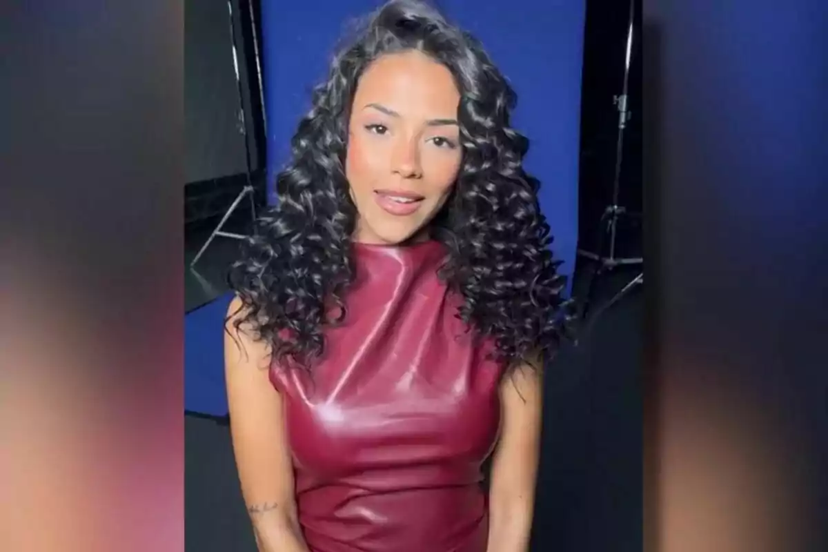 Jeimy Báez con cabello rizado y largo lleva un vestido de cuero rojo oscuro, posando frente a un fondo azul en un estudio fotográfico en GH DÚO.