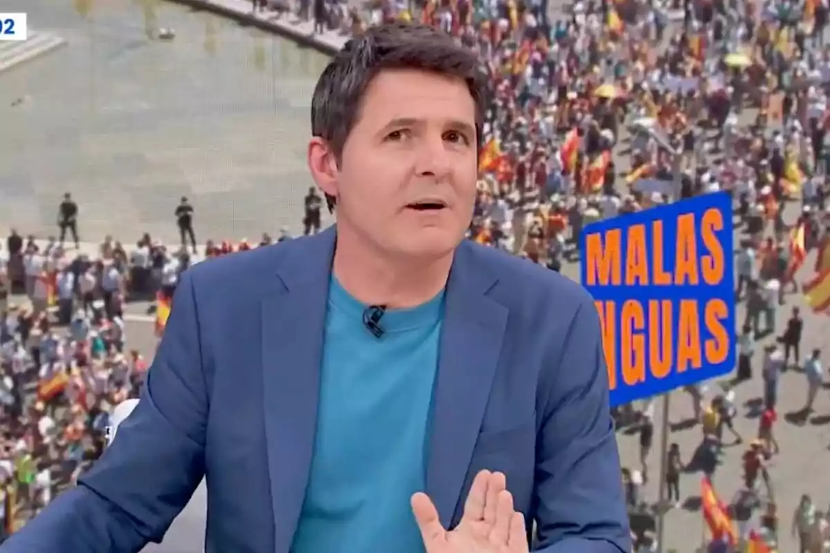 Jesús Cintora con chaqueta azul y camiseta turquesa está hablando frente a una multitud en una manifestación, con un cartel que dice "Malas Lenguas" al fondo.