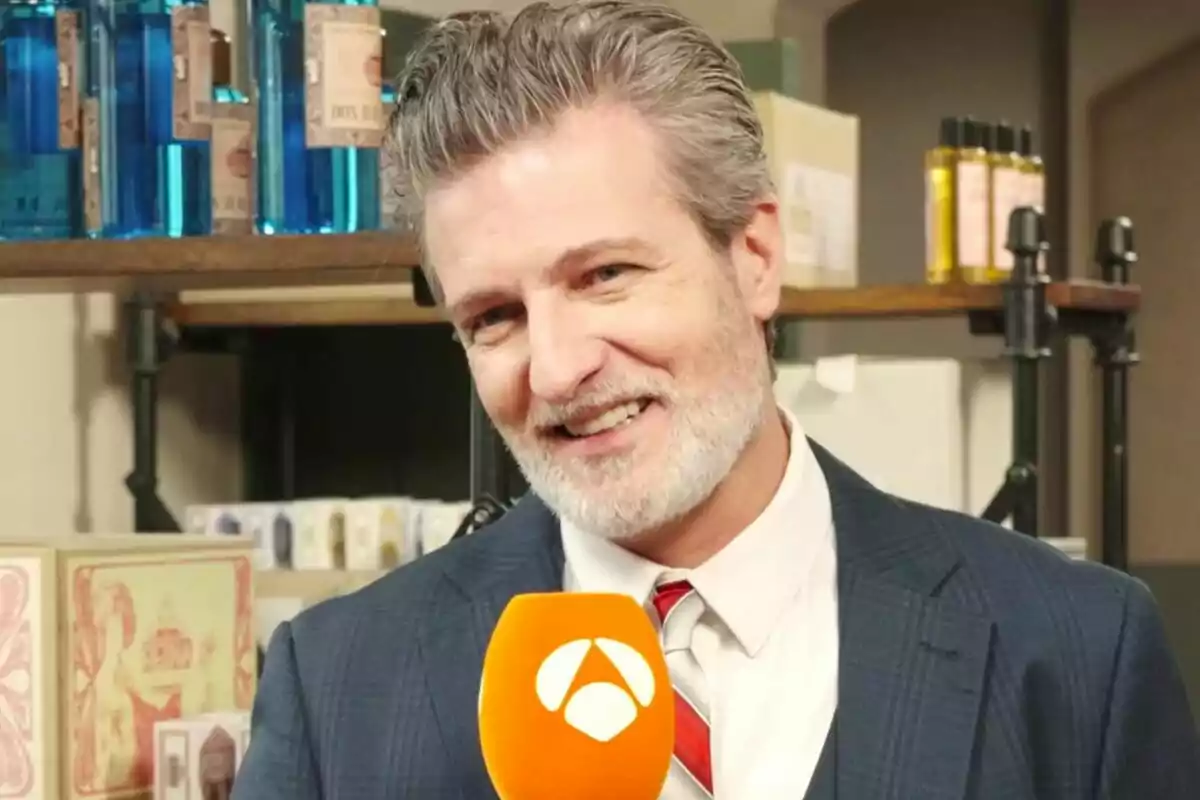 Jesús Olmedo con barba y cabello canoso, vestido con traje y corbata, sostiene un micrófono naranja con el logotipo de Antena 3, mientras sonríe frente a una estantería con productos en Sueños de Libertad.