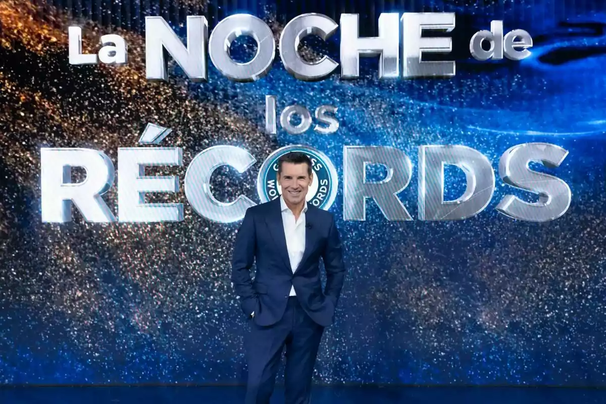Jesús Vázquez con traje azul oscuro sonríe frente a un cartel que dice Jesús Vázquez con traje azul oscuro sonríe frente a un cartel que dice