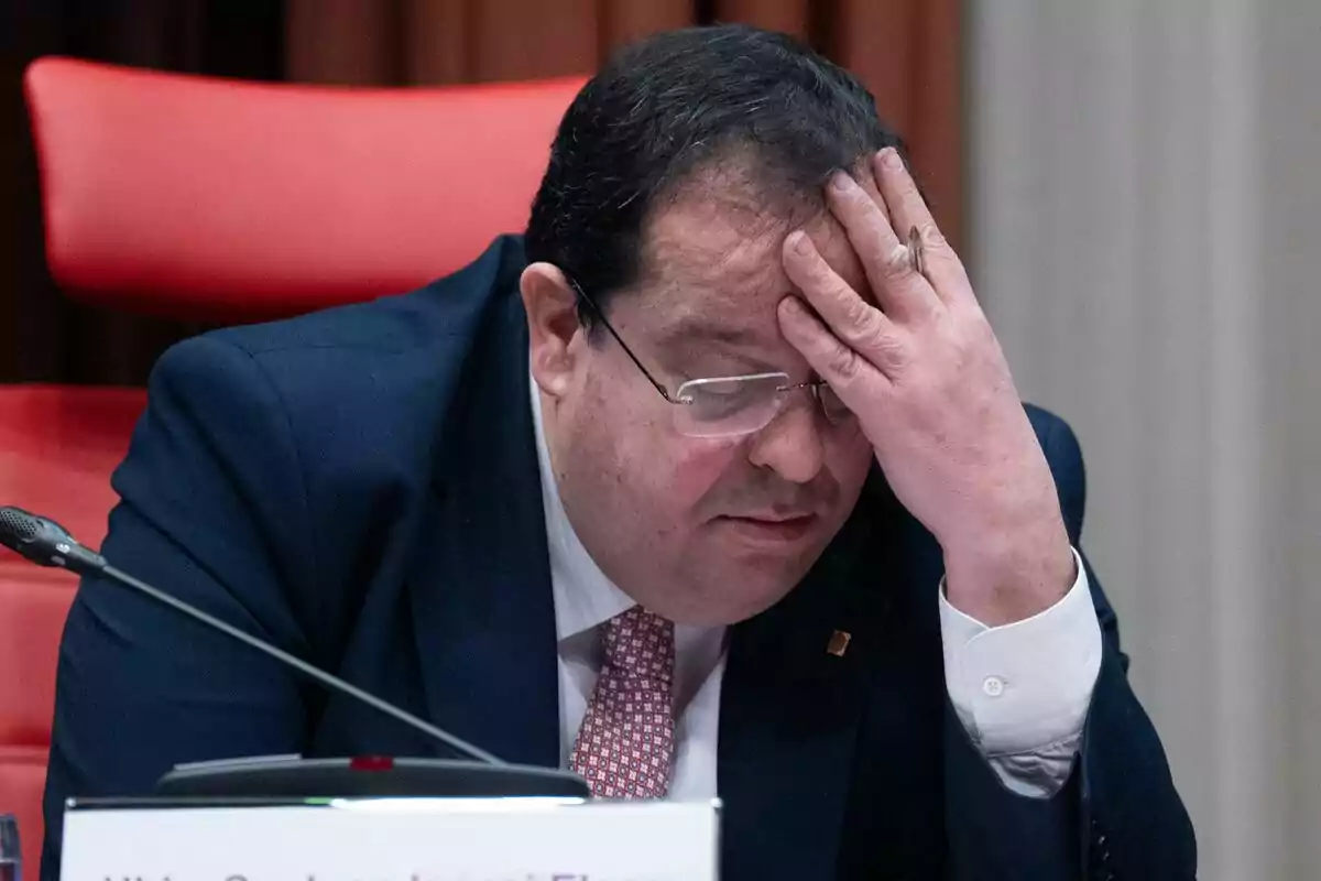 Joan Ignasi Elena con traje y corbata sentado en una silla roja, con la mano en la frente y expresión de preocupación.