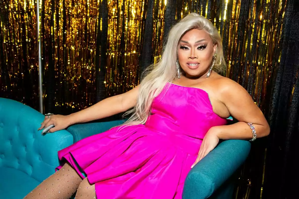 Jiggly Caliente con vestido rosa brillante y cabello rubio largo está sentada en un sofá azul con un fondo de cortinas doradas.