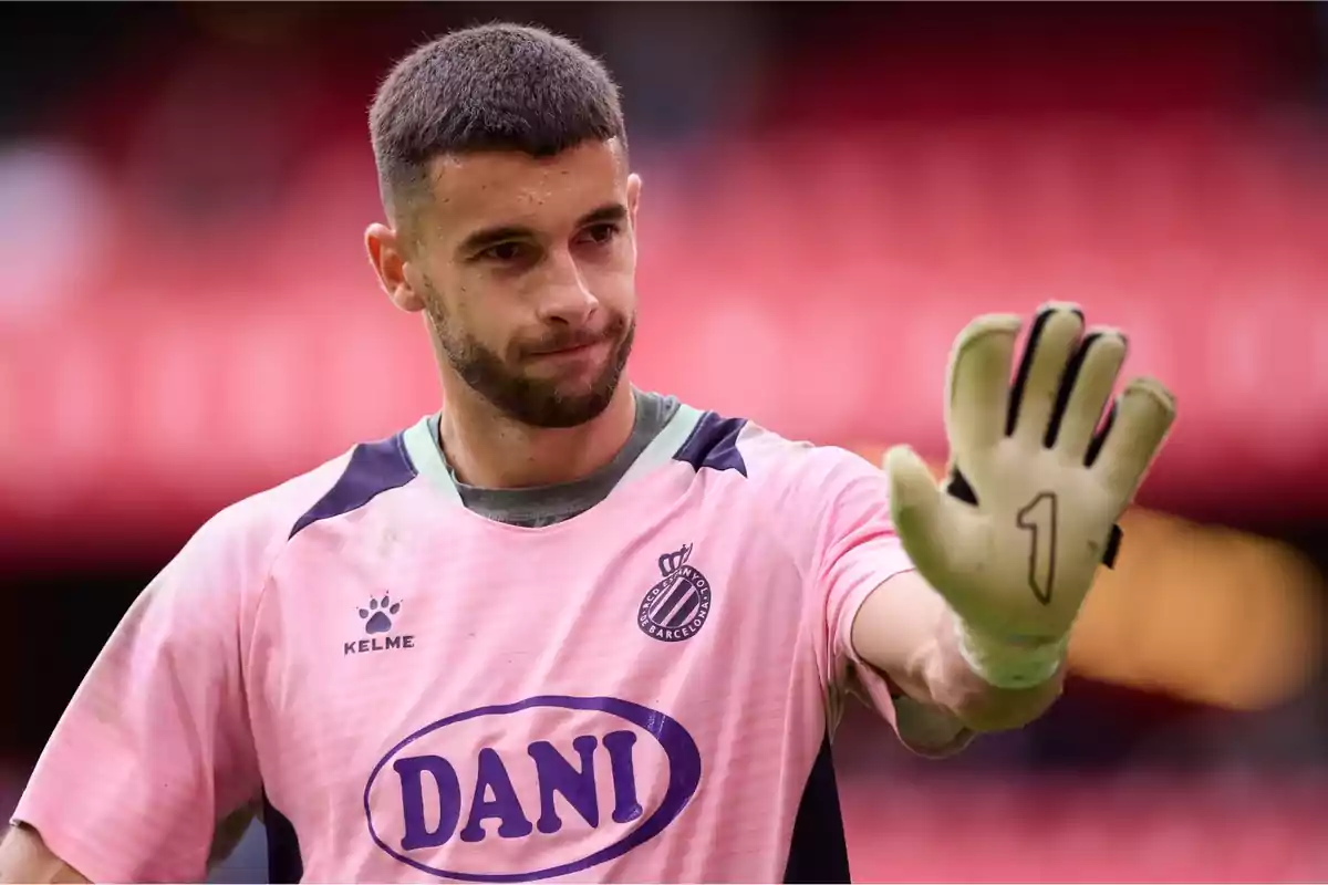 Un jugador de fútbol con uniforme rosa y guantes levanta la mano en señal de saludo. Un jugador de fútbol con uniforme rosa y guantes levanta la mano en señal de saludo.