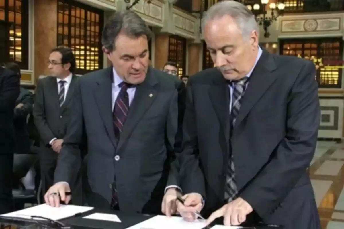 Dos hombres de traje están revisando documentos en una mesa en un entorno formal con otras personas al fondo.