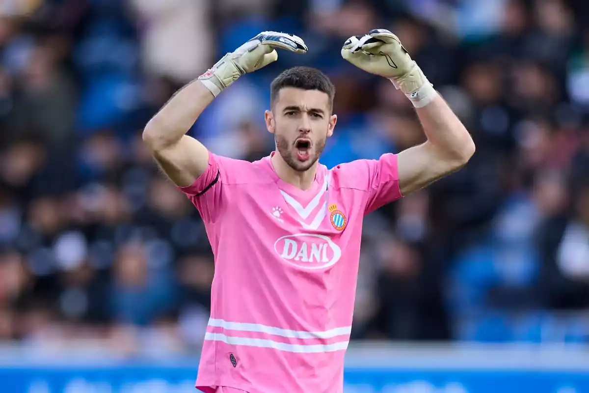 Un jugador de fútbol con uniforme rosa y guantes levanta los brazos en un estadio. Un jugador de fútbol con uniforme rosa y guantes levanta los brazos en un estadio.
