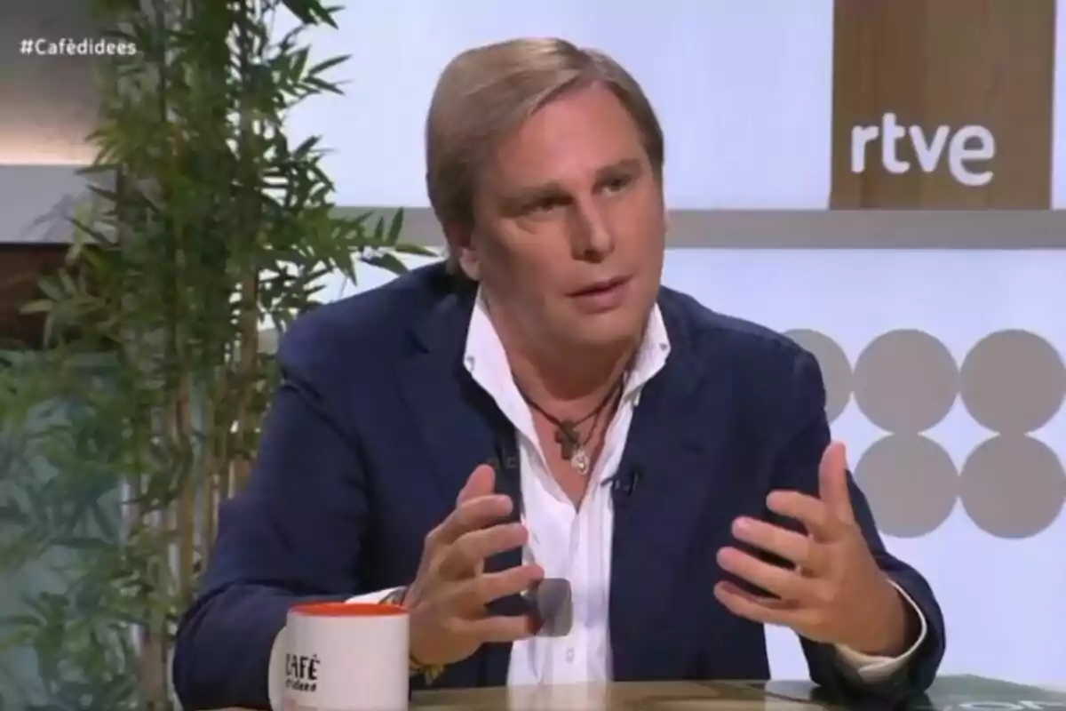 Un hombre con chaqueta azul y camisa blanca está hablando en un programa de televisión de RTVE, con una taza de café en la mesa y plantas en el fondo.