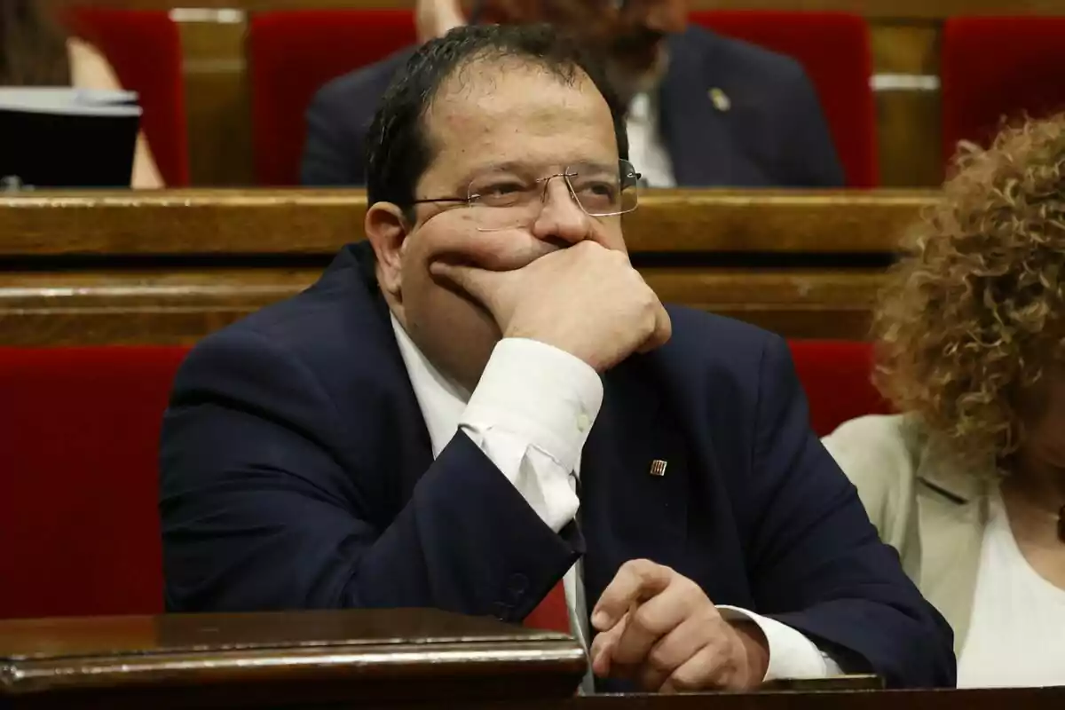 Joan Ignasi Elena está sentado en su escaño en el Parlament, apoyando su cabeza en su mano y mirando pensativo hacia adelante.