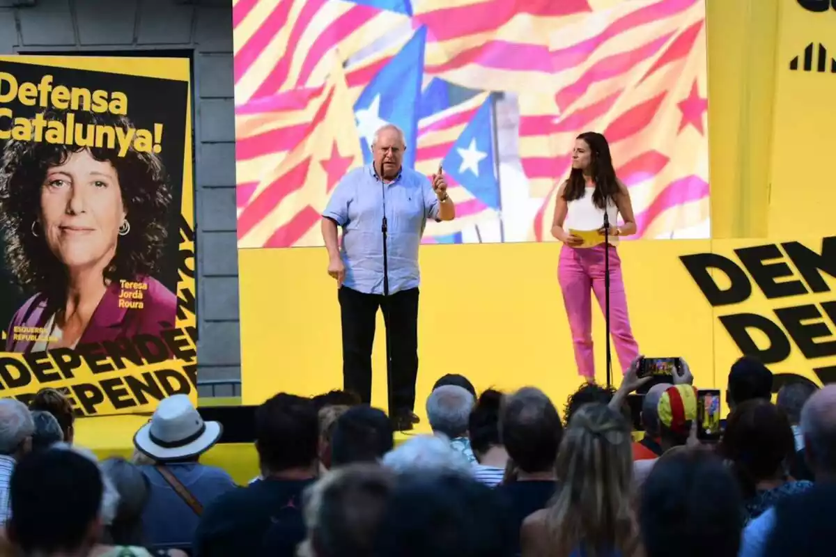 Plano general de Joan Queralt dando un mitin delante de decenas de personas. De fondo, una pantalla gigante amarilla con imágenes con banderas esteladas y un cartel electoral de Teresa Jordà