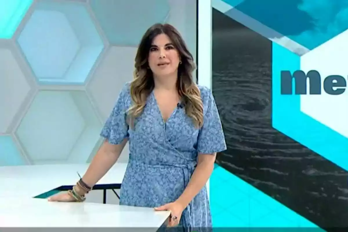 Joanna Ivars de pie en un estudio de televisión con un vestido azul y fondo geométrico. Joanna Ivars de pie en un estudio de televisión con un vestido azul y fondo geométrico.
