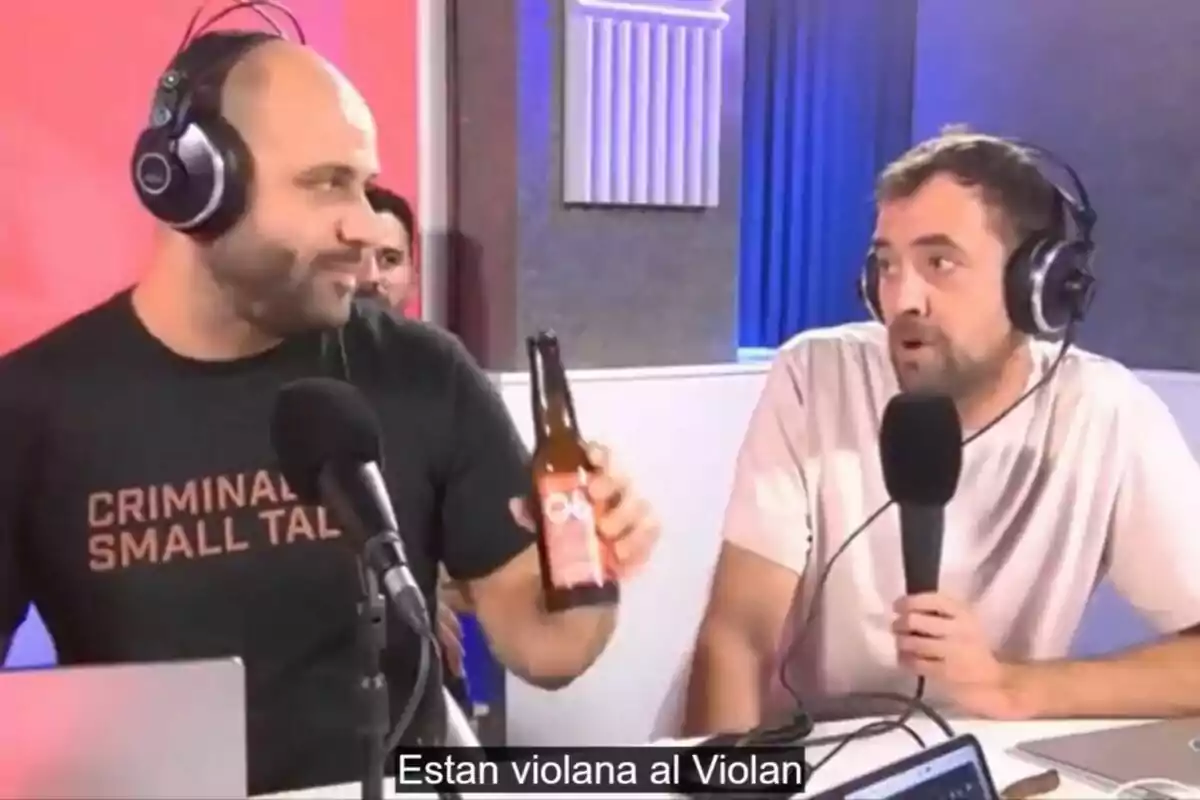 Dos personas con auriculares están en un estudio de grabación, una de ellas sostiene una botella y ambos parecen estar conversando frente a micrófonos.