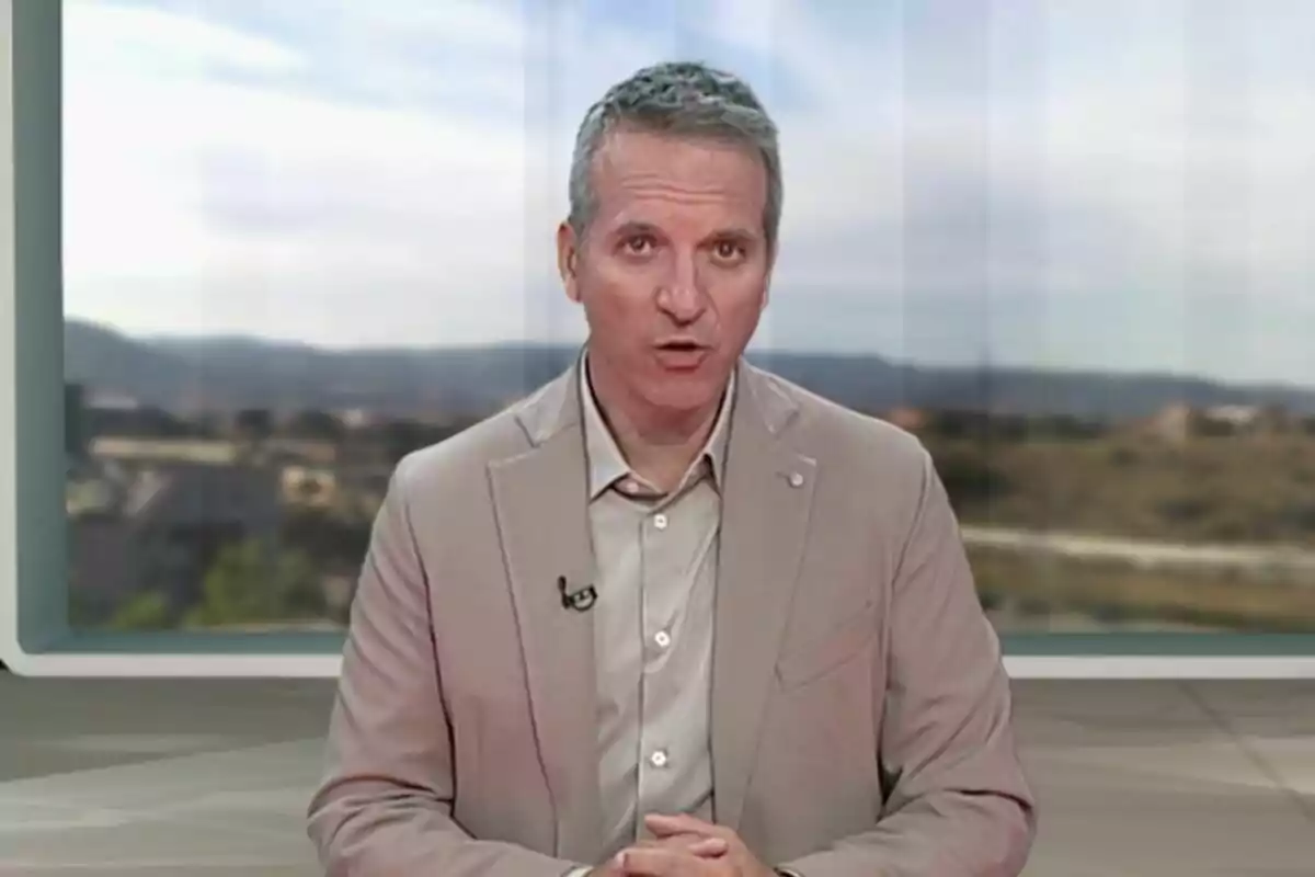 Plano medio de Jofre Janué vestido con saco beige hablando frente a una cámara en un estudio con fondo de paisaje difuminado.