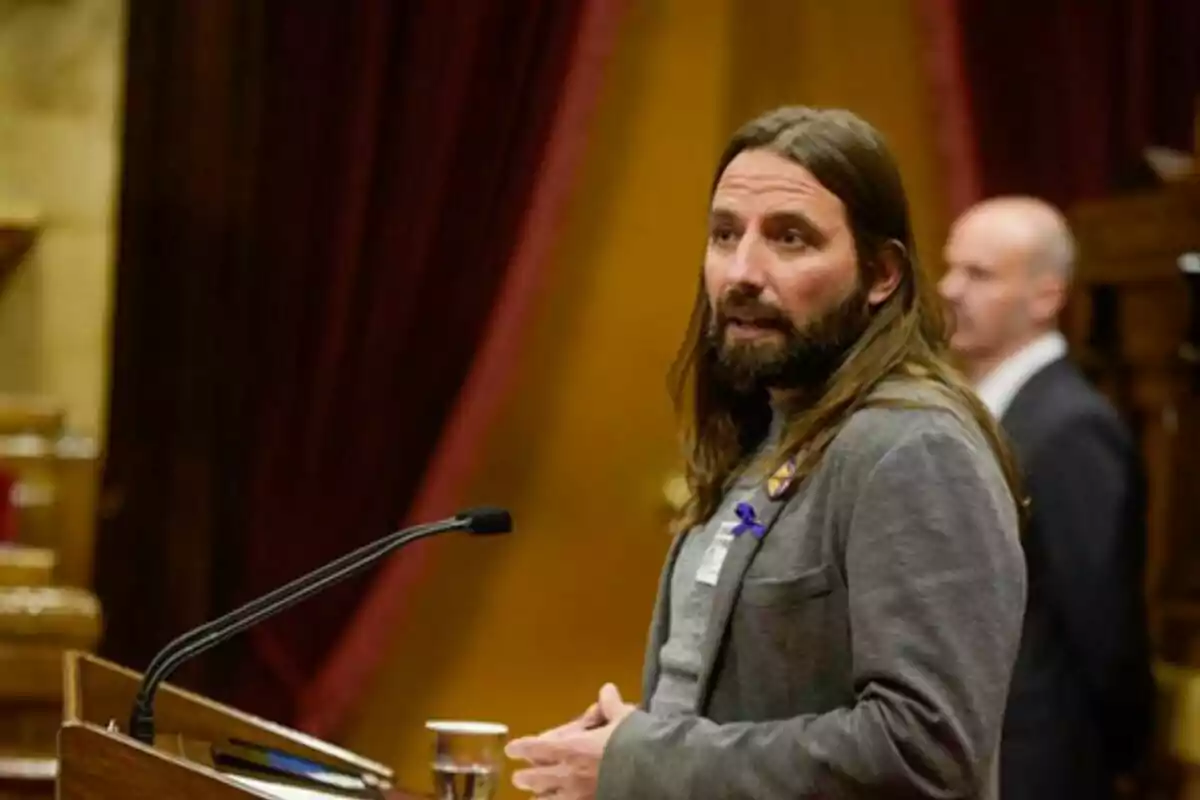 Un hombre con cabello largo y barba está hablando en un podio en un entorno formal con cortinas rojas al fondo. Un hombre con cabello largo y barba está hablando en un podio en un entorno formal con cortinas rojas al fondo.