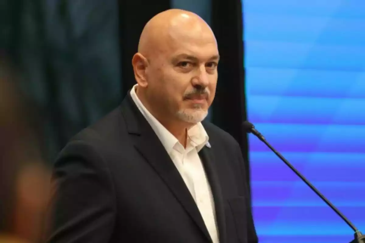 Hombre calvo con barba en traje oscuro hablando en un micrófono frente a un fondo azul.