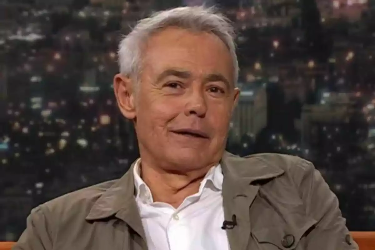 Jordi González de cabello canoso y corto con chaqueta beige y camisa blanca sentado en un set de televisión de Col·lapse con fondo de ciudad desenfocado